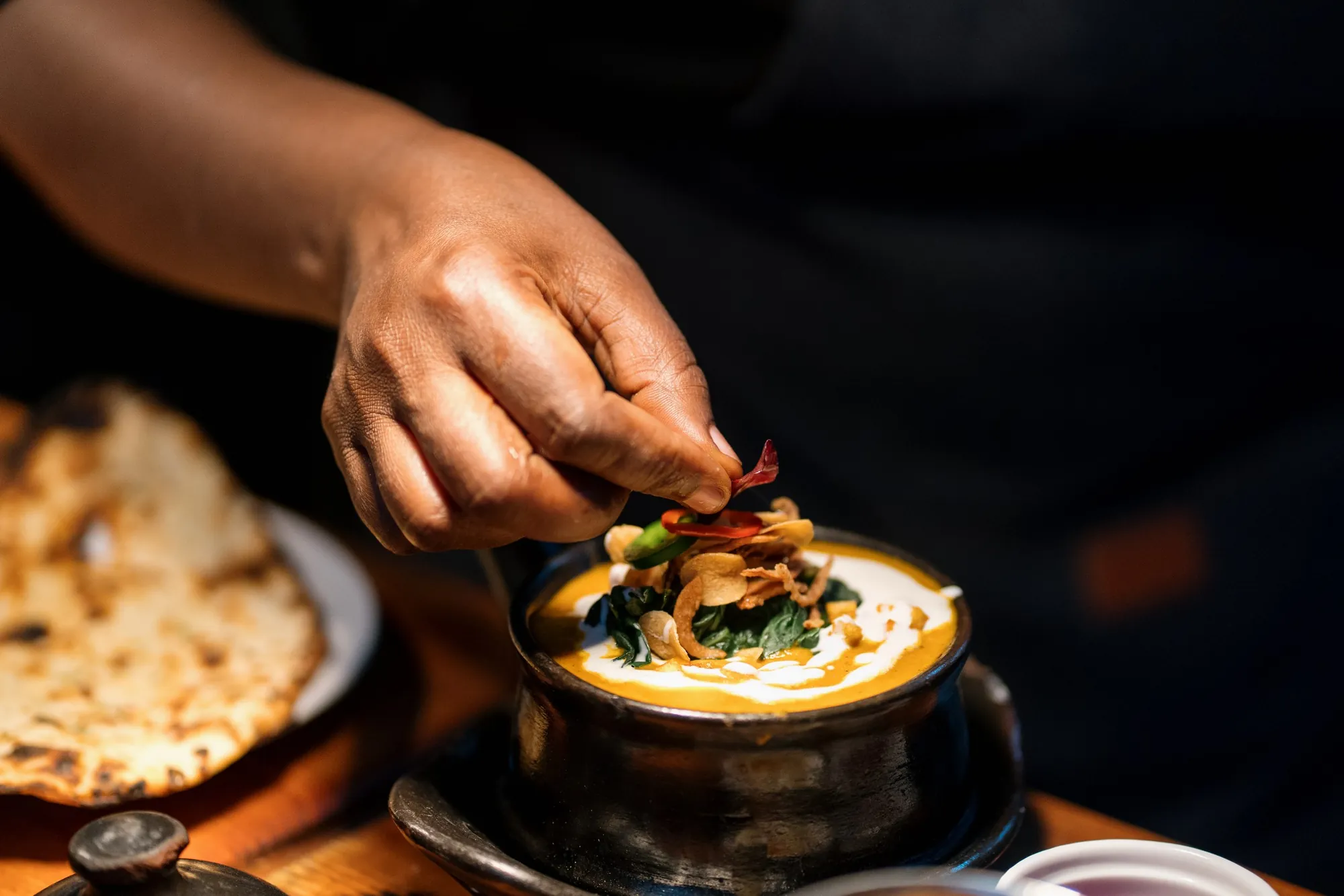 A chef garnishing a clay-pot dish beside fresh flatbread